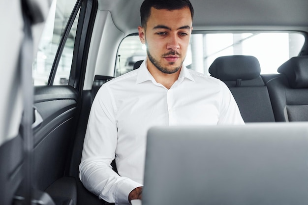 Works by using laptop Young man in white shirt is sitting inside of a modern new automobile