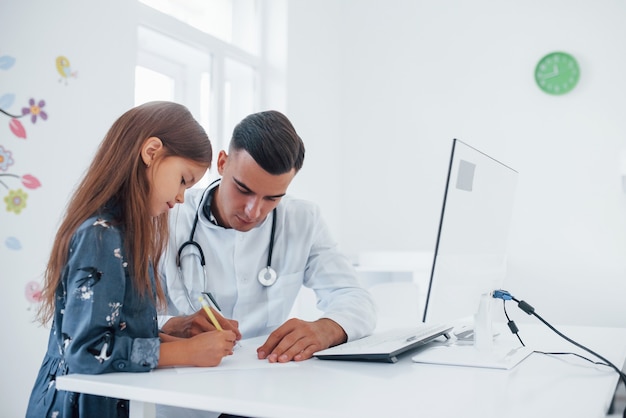 Works with documents. Young pediatrician works with little female visitor in the clinic.