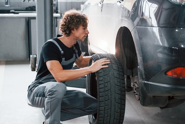 Works with tire Adult man in grey colored uniform is in the automobile salon