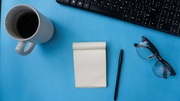 workspace desk with notebooks keyboard pens glasses and coffee on blue background