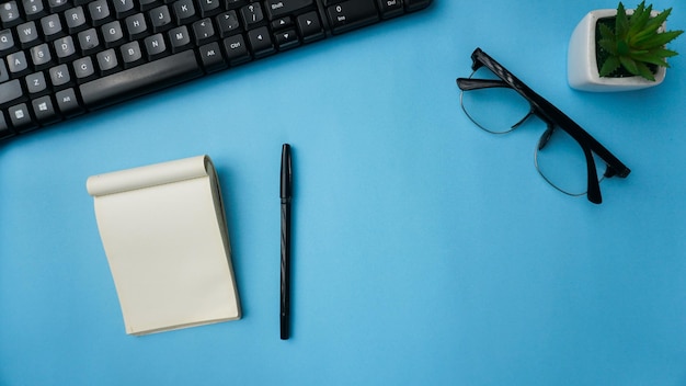 workspace desk with notebooks keyboard pens glasses plants on blue background