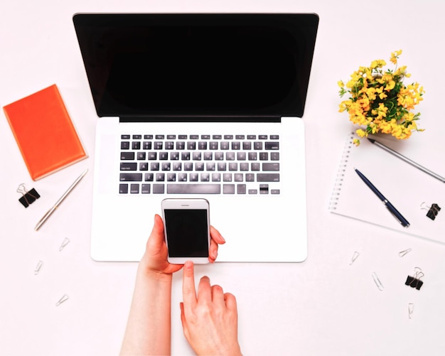 Workspace with female hand holding mobile phone and laptop keyboard, and yellow flowers on the white background. Flat lay, top view