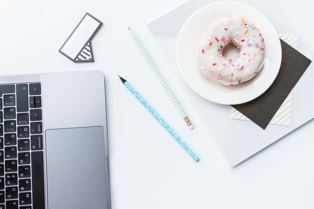 Area di lavoro con laptop, matita, quaderno e ciambella su sfondo bianco