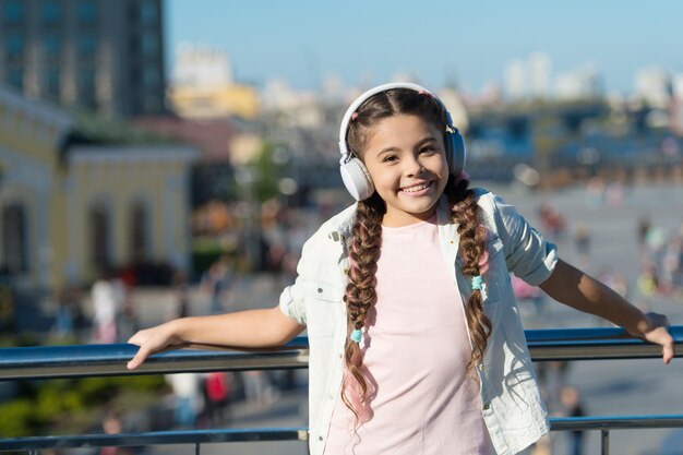 ヘッドホンの向こうの世界はカオスすぎる。都市の屋外でヘッドフォンを着ている少女。耳のステレオ ヘッドフォンで音楽を聴いているかわいい子。現代のヘッドフォンを身に着けているファッショナブルな小さな子供。