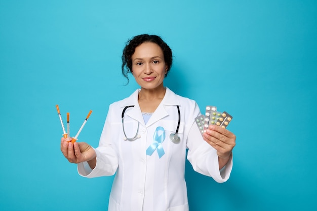 Giornata mondiale del diabete. 14 novembre. bella dottoressa in abito medico bianco che indossa un nastro di consapevolezza blu tiene siringhe da insulina e compresse farmaceutiche in blister, sorride guardando la fotocamera.