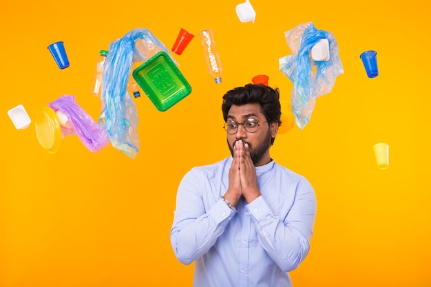 World Environment Day, plastic recycling problem and environmental disaster concept - Scared Indian man is standing under trash and looking side on yellow background