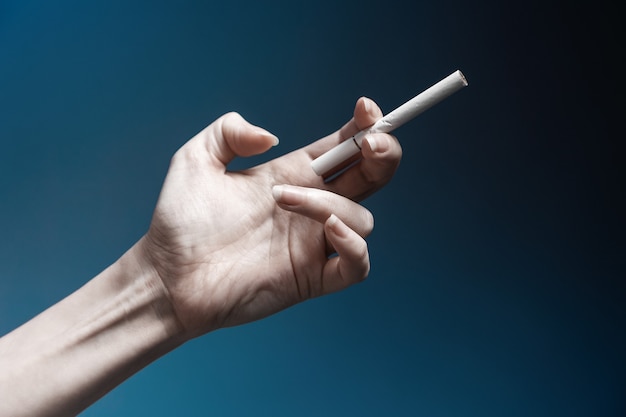 Photo world no tabacco day. a pale, crooked female hand, close-up, holding a new cigarette. dark blue background. the concept of nicotine addiction.