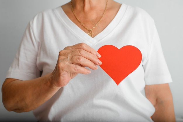 World stroke day, heart disease concept. Close-up of elderly woman holding red paper heart with wrinkled hands on background of white t-shirts. Selective focus on the heart