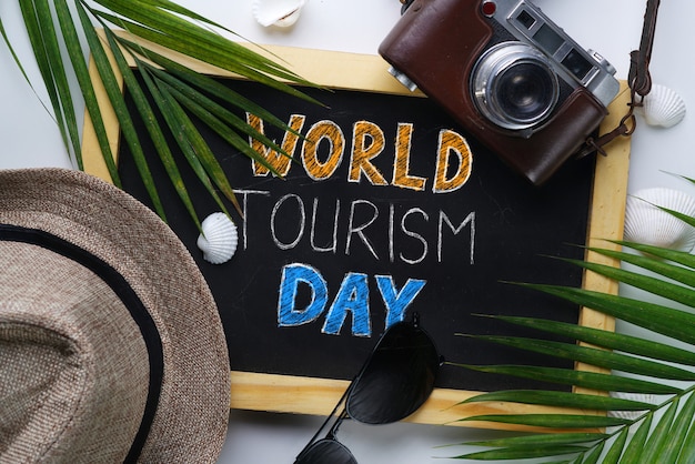 Photo world tourism day typography. sunglasses, fedora hat, palm leaf, camera, sea shells and blackboard