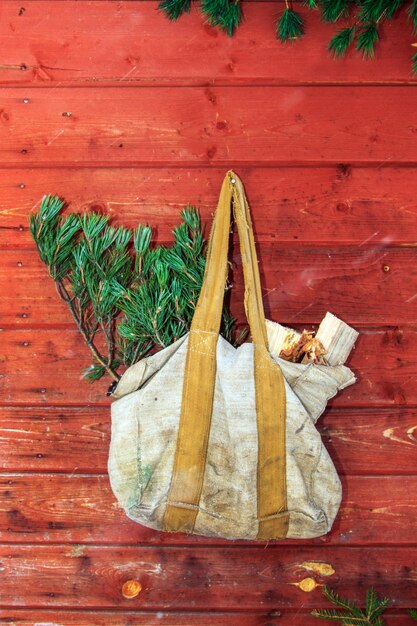 Фото Изношенная холстовая сумка с бревнами и еловыми ветвями висит на красной подлинной деревянной стене
