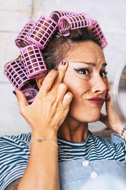 Worried adult young caucasian female check wrinkles and aging time effect on her face skin beautiful funny woman with make up and curlers on hair at home in getting ready activity