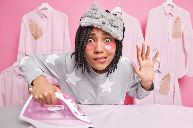 Photo worried afro american woman with dreadlocks bites lips keeps palm raised looks attentively at camera strokes clothes uses electric iron wears sleepamask soft pajama