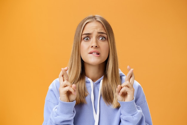 Worried anxious hopeful young woman with fair hair in stylish over-sized hoodie biting lower lip frowning concerned crossing fingers for good luck praying for dream come true against orange wall