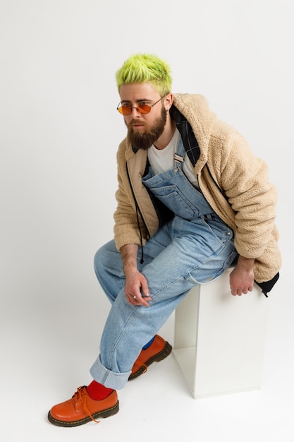 Worried attractive man dresses stylish attire and glasses looking away with sad facial expression upset male model sitting on white cube Indoor studio shot isolated over gray background