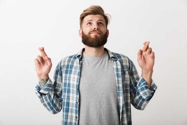 Worried bearded man dressed in plaid shirt standing isolated, holding fingers crossed for good luck