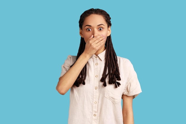 Worried beautiful woman with dreadlocks wants to scream, covers mouth with palm, stares at something terrible, has eyes popped out, wearing white shirt. Indoor studio shot isolated on blue background.