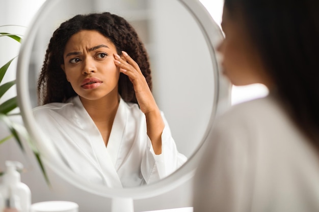 Обеспокоенная черная женщина смотрит в зеркало и трогает морщины на лице
