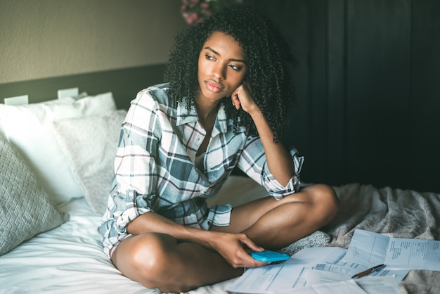 Worried black woman with bills and finance problems on bed