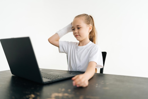 Worried blonde little girl put broken hand wrapped in white\
plaster bandage on head thoughtful looking to laptop screen sitting\
at table on white background