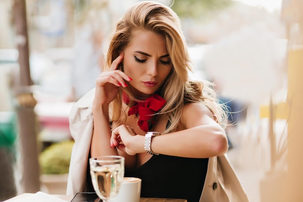 Worried blonde woman looking at wristwatch while enjoying coffee in outdoor restaurant. Unpleased tanned girl with nude make-up waiting friend in cafe drinking water on blur background.