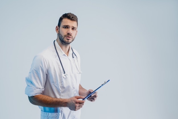 Worried caucasian male doctor with stethoscope and clipboard. Young bearded man wearing white coat. Isolated on gray background with turquoise light. Studio shoot. Copy space.