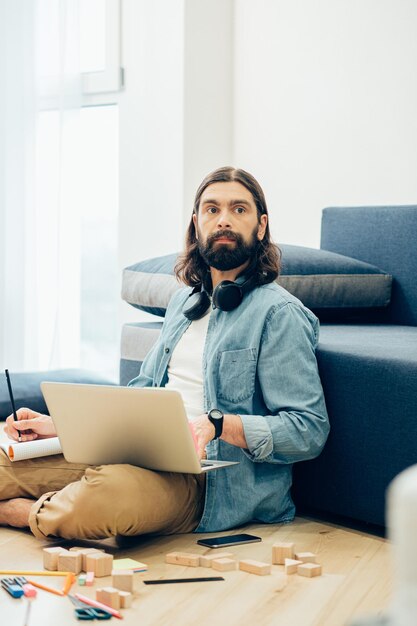 Worried Caucasian young man sitting on the floor with a laptop and looking into the distance