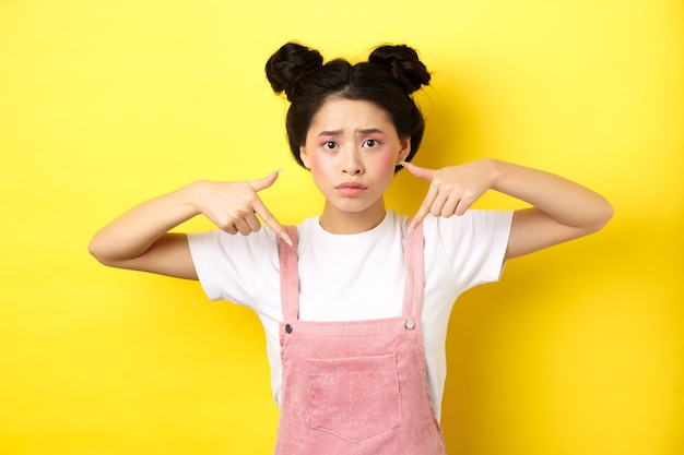 Worried and confused asian girl with glamour makeup, pointing fingers down and look sad, standing in summer clothes against pink.