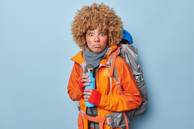 Worried curly female explorer has frozen face holds thermos\
with hot beverage carries heavy rucksack wears orange windbreaker\
and scarf discovers something new isolated over blue\
background