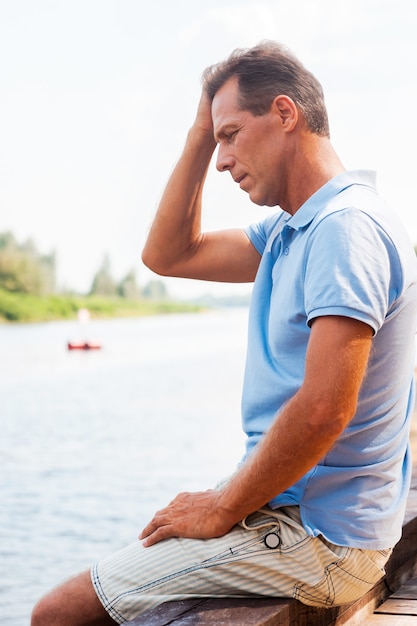 Photo worried and depressed man. side view of depressed mature man touching head with hand and looking down while sitting at the quayside