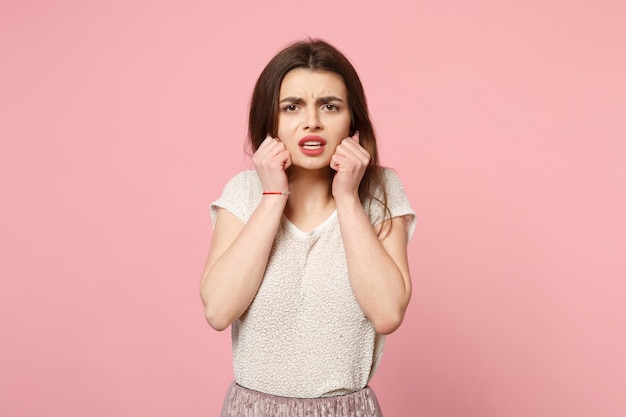 Foto giovane donna dispiaciuta preoccupata in abiti leggeri casual in posa isolata su sfondo rosa ritratto in studio. persone sincere emozioni concetto di stile di vita. mock up spazio di copia. stringere i pugni vicino alle guance.