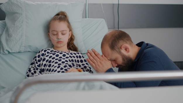 Worried father praying for sick girl daughter health recovery\
after suffering medical surgery in hospital ward. little child with\
oxygen nasal tube sleeping during sickness examination