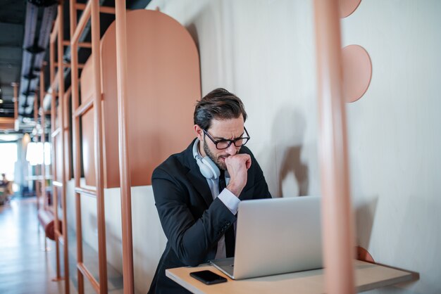 Worried handsome caucasian bearded businessman in suit and with eyeglasses sitting at table with hand on chin and checking on reports.