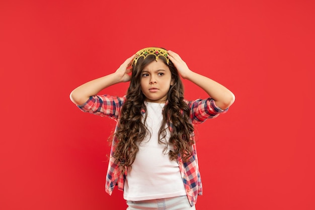Worried kid with curly hair in princess crown on red background winner
