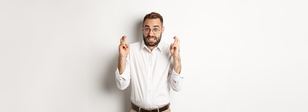 Worried man making a wish cross fingers and hope for relish standing over white background