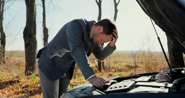 Worried man in a safety vest holding his head by hand standing near his broken car with raised hood on the road. Concept road accident. Help repair.