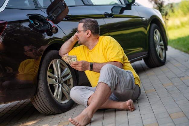 Photo worried man sitting on the pavement next to a car near an open fuel tank fuel price rise concept