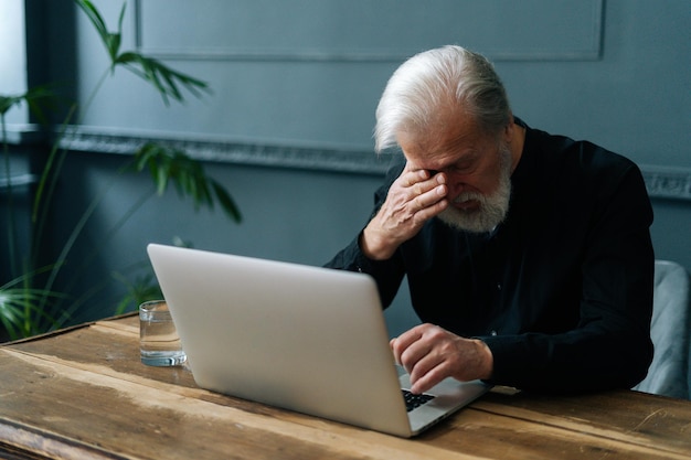 Photo worried mature gray-haired grandfather grieving, received bad message notification, sitting alone at home. depressed bearded senior aged man crying looking at laptop computer screen in dark room.