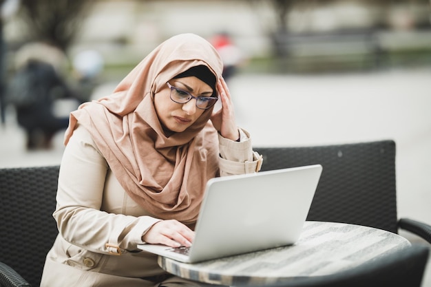 Worried Muslim business woman wearing a hijab and using her laptop while sitting in urban cafe.