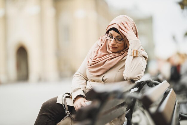 Worried Muslim woman wearing a hijab and pensive sitting on the bench in urban environment.