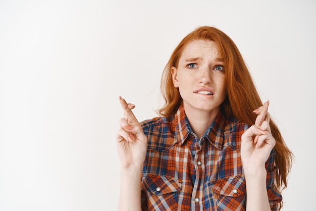 写真 心配している赤毛の女の子が唇を噛み、白い背景の上に立って願い事をしたり、結果を期待して嘆願したりしながら、希望のクロスフィンガーで見上げる