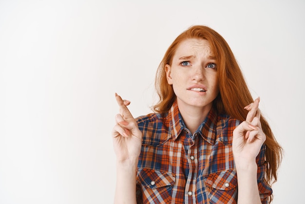 Photo worried redhead girl biting lip and looking up with hope cross fingers while making a wish or pleading anticipating results standing over white background