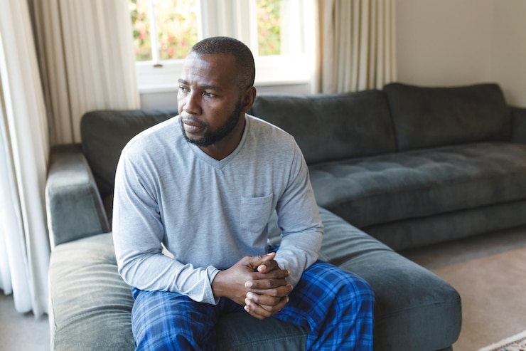 Photo worried, sad african american man thinking, sitting on couch looking out of window in living room