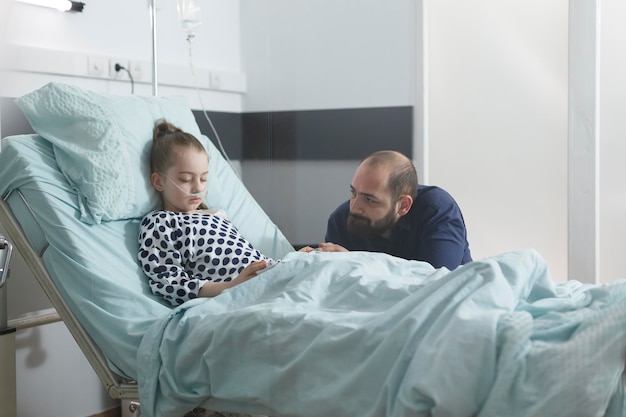 Photo worried sad uneasy father watching sick little daughter sleeping in hospital bed while in pediatric clinic patient room. under treatment ill child resting while attentive parent taking care of her.