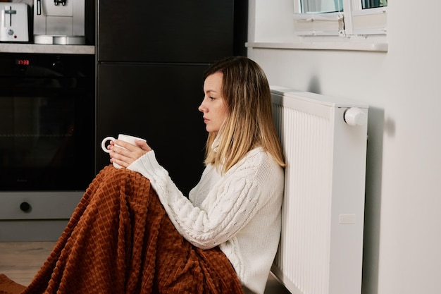 写真 心配している悲しい女性は、お茶を飲みながらラジエーターを加熱する近くの毛布の下に座っています