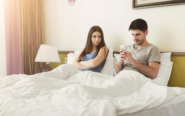 Worried woman looking at husband with smartphone addiction while couple sitting on bed