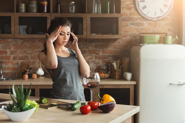 Worried woman talking on phone and drinking wine while cooking healthy food in the loft kitchen at home on sunny day. Problems and bad news concept, copy space