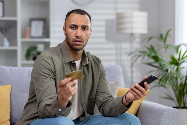Worried young african american man sitting on couch at home and looking upset at camera holding