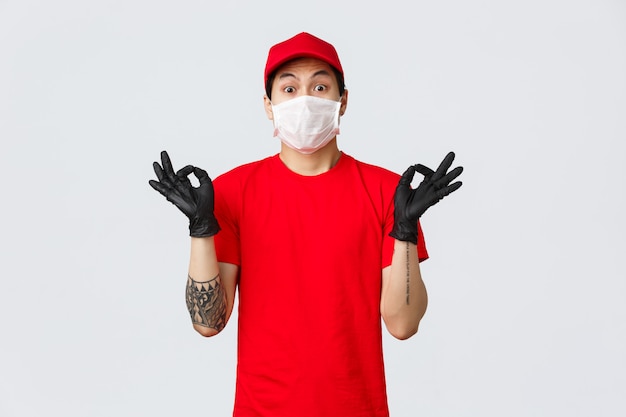 Потревоженный молодой азиатский работник доставляющий покупки на дом пробуя успокоить слабонервного клиента, показывая жест Дзэн, нося медицинскую маску и защитные перчатки, форму компании. Курьер делает хорошо знаком обеспечить лучшее качество