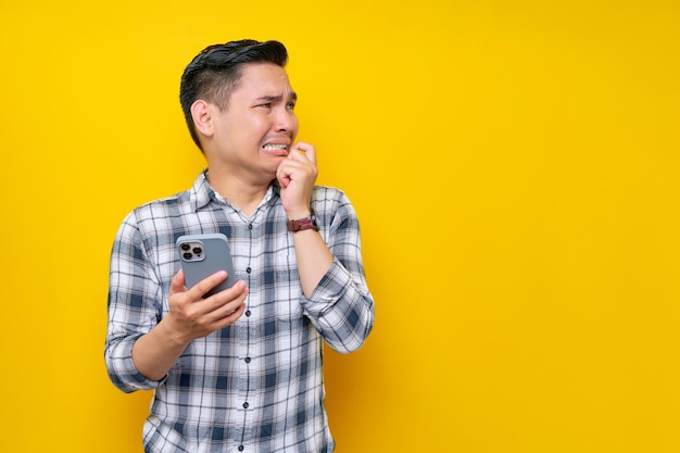 Worried young asian man wearing a white checkered shirt holding smartphone and biting his nails looking side with a frustrated expression isolated over yellow background people lifestyle concept