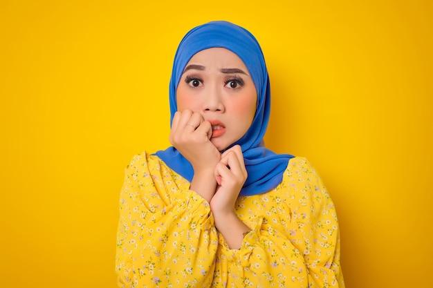 Worried young Asian woman in casual dress biting nails looks stressed and nervous isolated on yellow background